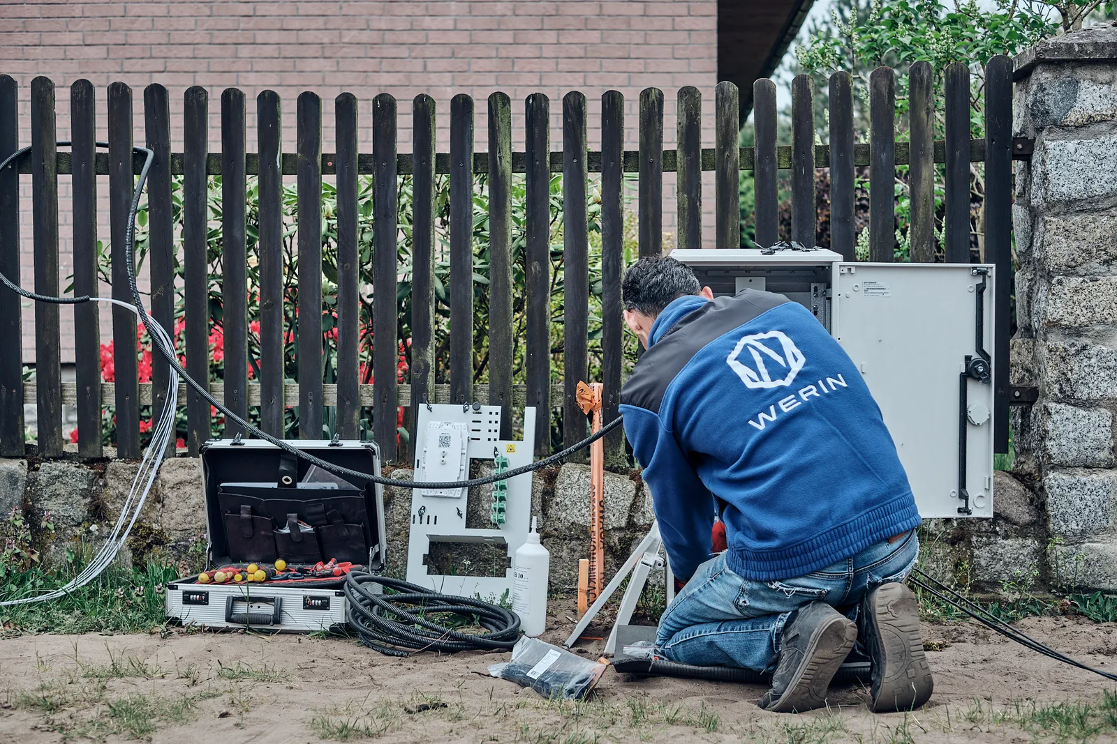 Technik WERIN klęczy przy otwartej szafie dostępowej pod płotem domu, obok walizka z narzędziami i zwój kabla