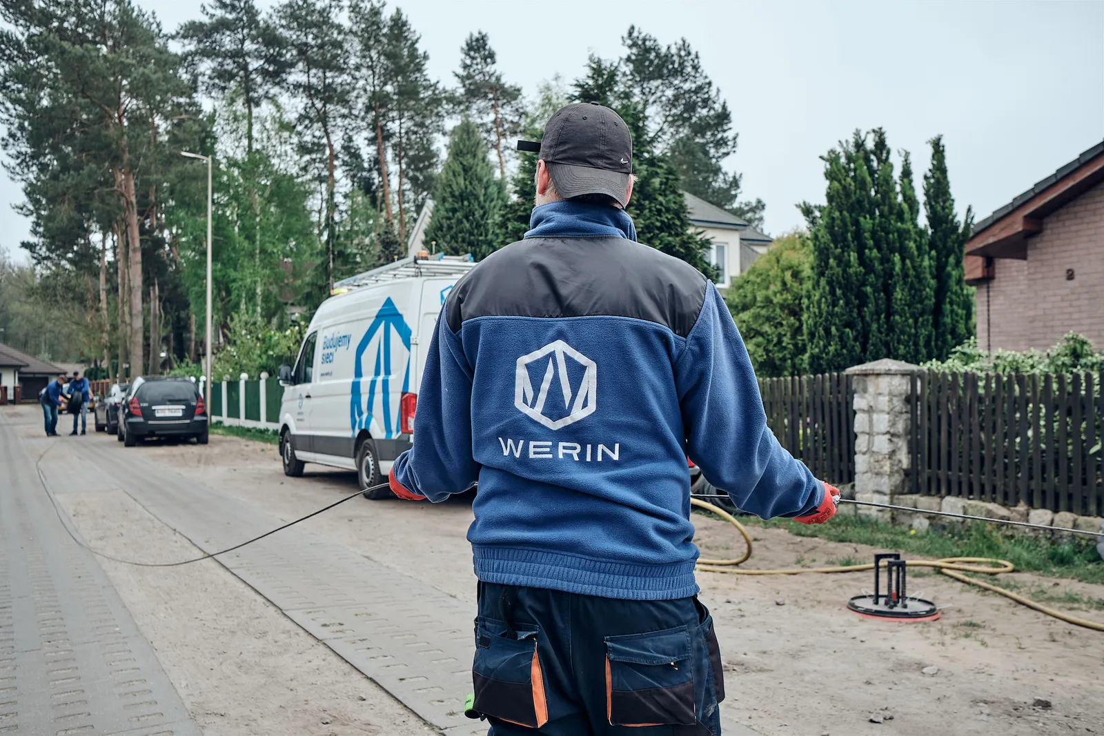 Brygadzista WERIN tyłem na ulicy osiedla domów, w tle firmowy bus z napisem Budujemy sieci